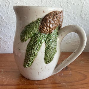 May include: Cream ceramic mug featuring a brown pine cone and green pine needles. The mug has a speckled finish and a curved handle. The pine cone and needles are raised, adding texture to the design. A rustic style mug.