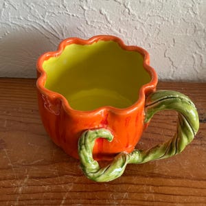 Op de afbeelding: Een keramische mok in de vorm van een pompoen met een glanzende oranje buitenkant en een felgele binnenkant. De mok is voorzien van een gebeeldhouwd, gedraaid groen rankhandvat dat aan de zijkant uit de mok steekt.