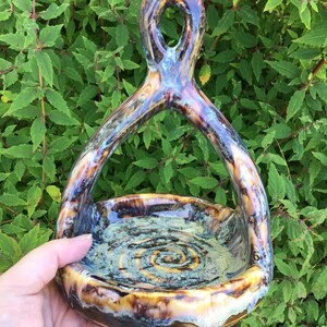 Puede incluir: Un cuenco de cerámica artesanal con un asa única, sostenido en una mano. El cuenco presenta un diseño en espiral en el centro y está esmaltado con una mezcla de tonos marrones, azules y amarillos. El asa tiene una abertura ovalada en la parte superior.