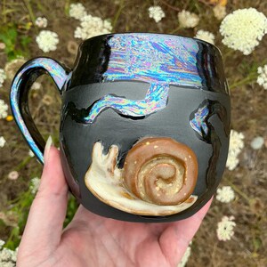 May include: A black ceramic mug featuring a sculpted, light brown snail on the front. The mug has a glossy, iridescent finish on the handle and upper rim, with abstract, shimmering blue and purple streaks across the dark matte surface.