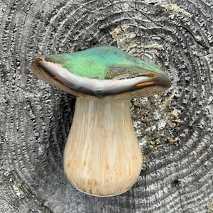 May include: A ceramic mushroom figurine with a green and bronze cap and a cream-colored stem. The mushroom has a glossy finish and is set against a textured, gray background.
