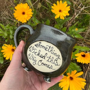 May include: A dark gray ceramic cauldron mug with a rounded body, a curved handle, and three small feet. The front features a cream-colored circular patch with the black handwritten text, "Something Wicked this Way Comes" in cursive script.