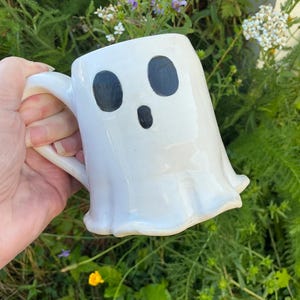 May include: A white ceramic mug shaped like a ghost with a smooth, glossy finish. The mug features two large, oval-shaped black eyes and a smaller, vertically oriented black oval mouth painted on the front surface, with a curved handle on the side.