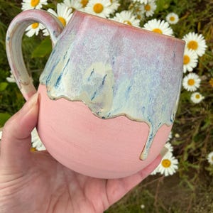 May include: A ceramic mug with a matte pink base and a glossy, dripping glaze in shades of light blue, cream, and soft pink. The mug features a curved handle and a rounded body, with the glaze creating a distinct, uneven drip pattern that cascades down from the rim toward the center of the vessel.
