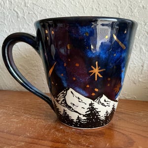 May include: A ceramic mug with a dark blue, starry night sky design. Gold accents depict stars and shooting stars. The bottom of the mug features a black and white mountain range and pine trees.