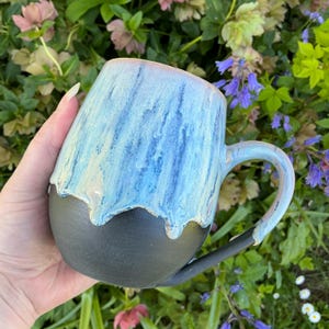 May include: A handmade ceramic mug with a unique design. The mug features a black base, transitioning to a light blue and cream-colored top with a dripped glaze effect. The handle is also light blue. A beautiful piece for coffee or tea.
