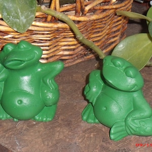 Puede incluir: Dos figuritas de rana verde, una sentada con las piernas cruzadas y la otra de pie con los brazos extendidos. Las ranas están hechas de un material liso y brillante.