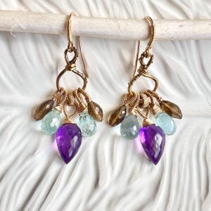 May include: A pair of gold-toned wire-wrapped dangle earrings featuring clusters of faceted teardrop gemstones. Each earring includes a central purple amethyst, two light blue aquamarine stones, and two brown smoky quartz beads suspended from a hook.