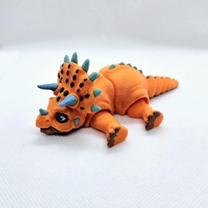 May include: An orange triceratops figurine with blue horns and spikes. The dinosaur has black spots on its head and is lying down. The figurine is made of a smooth material and has detailed feet.
