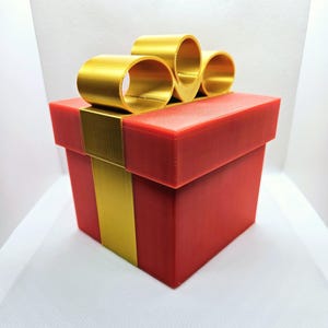 Puede incluir: Una caja de regalo roja con una cinta y un lazo dorados. La caja está envuelta en papel rojo con una cinta y un lazo dorados.