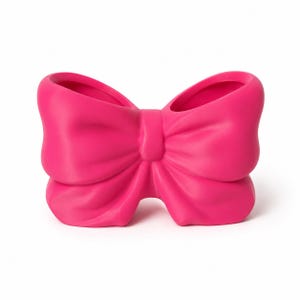 Puede incluir: Un recipiente decorativo de color rosa intenso con forma de lazo grande. El objeto presenta un acabado mate y liso con dos compartimentos abiertos distintos en la parte superior, creando una estructura hueca que imita un lazo atado.