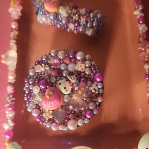 May include: Pink and purple resin tray with a pink and purple Hello Kitty design. The tray is decorated with rhinestones, pearls, and glitter. The tray has a small compartment for a lighter and a larger compartment for a small container.