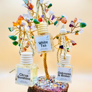 May include: A decorative tree sculpture with gemstone leaves and three hanging fragrance bottles. The bottles are labelled "White Tea", "Citrus Sunshine", and "Oakmoss & Amber". The tree trunk is gold-coloured.