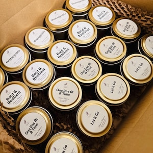 May include: A collection of round metal candle tins with gold lids arranged in a cardboard box. Each lid features a white circular label with black text reading Unstoppable, Bold & Resilient, One Day At A Time, or Let It Go, plus Made in California.