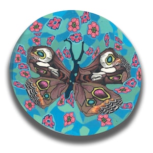 May include: A round, decorative object featuring a detailed butterfly design. The butterfly has brown wings with yellow, pink, and blue accents, surrounded by pink flowers on a turquoise and blue background. The object is likely a coaster or decorative piece.