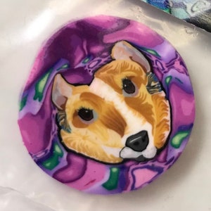 May include: A round, handcrafted polymer clay pendant featuring a detailed portrait of a dog's face. The dog has tan and white fur with black eyes and nose, set against a swirling purple, pink, and green background.