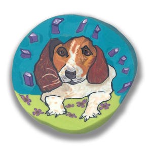 Könnte beinhalten: Ein rundes, bemaltes Keramikstück mit einem Basset Hound. Der Hund hat braunes und weißes Fell mit langen, hängenden Ohren. Der Hintergrund ist türkis mit lila geometrischen Formen, und der Vordergrund ist grün mit lila Blumen.