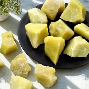 Puede incluir: Una colección de piedras de jade en bruto, de color verde claro, algunas sobre un plato negro, otras dispersas. Las piedras tienen una textura rugosa y natural y diferentes tonos de verde y amarillo. Un pequeño jarrón blanco con vegetación en el fondo.