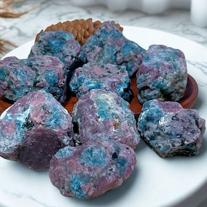 Könnte beinhalten: Eine Sammlung roher, ungeschliffener Rubin-in-Zoisit-Steine. Die Steine zeigen eine Mischung aus tiefen Rosa-, Blau- und Grüntönen. Sie sind auf einem Holztablett und einem weißen Teller angeordnet und zeigen ihre natürliche, unpolierte Textur.