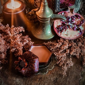 May include: A still-life arrangement featuring a lit candle, a brass coffee pot, and a halved pomegranate. A small, ornate container and dried flowers are also present. The scene is set on a wooden surface, with warm, inviting tones.