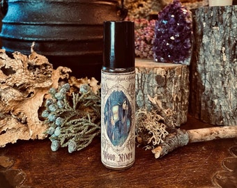 May include: A brown glass roll-on bottle with a black cap. The bottle has a label with a drawing of a woman in a forest with the text "Goddess Magic Essences Wood Nymph Botanical Ritual Oil".