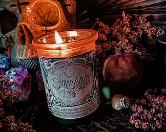 May include: A lit candle in a glass jar with the word "Samhain" on the label. The candle sits on a dark surface with a skull, crystals, and dried botanicals. The label also includes the text "Moon Goddess Magick Apothecary".