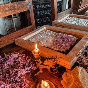 May include: Two wooden frames with white paper and lilac flowers inside, a lit candle on a dragon-shaped candlestick, and a lit candle on an orange slice. The frames are on a wooden table with lilac flowers and a piece of beeswax.