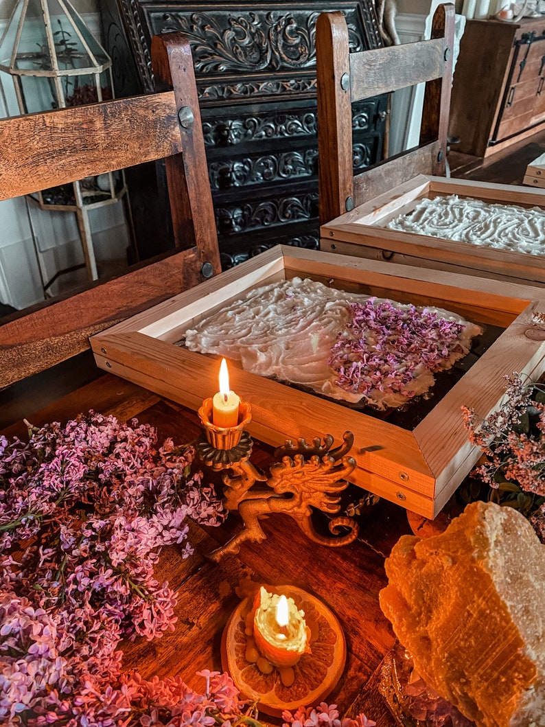 May include: Two wooden frames with white paper and lilac flowers inside, a lit candle on a dragon-shaped candlestick, and a lit candle on an orange slice. The frames are on a wooden table with lilac flowers and a piece of beeswax.