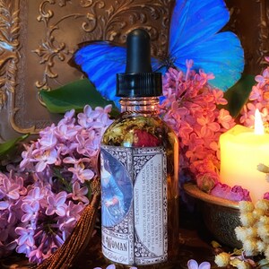 May include: A glass bottle with a dropper filled with a clear liquid and dried flowers. The bottle has a label with the text "The Enchantment" and "Woman Body Oil". The bottle is surrounded by purple and pink flowers.