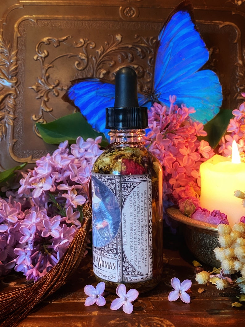 May include: A glass bottle with a dropper filled with a clear liquid and dried flowers. The bottle has a label with the text "The Enchantment" and "Woman Body Oil". The bottle is surrounded by purple and pink flowers.