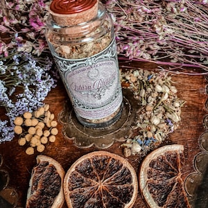 May include: A glass jar with a cork lid and a label that reads "Ostara Blessing" filled with dried herbs and flowers. The jar is sitting on a wooden surface with dried orange slices and dried flowers.