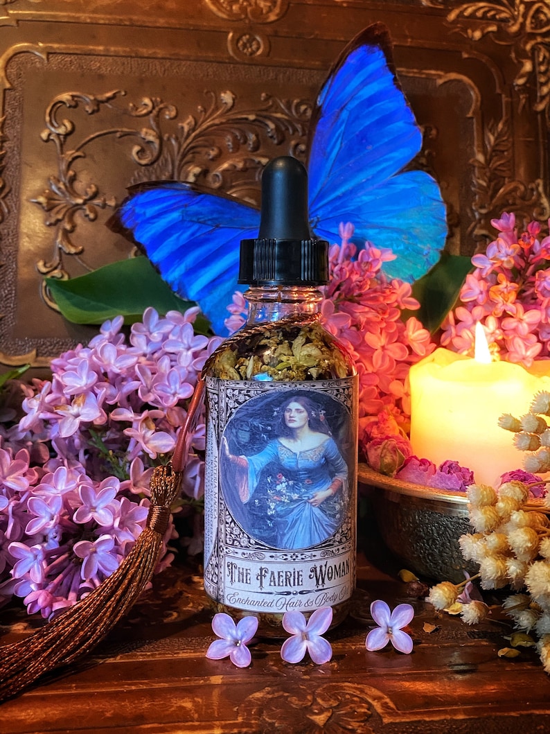 May include: A glass bottle with a dropper top and a label that reads "The Faerie Woman Enchanted Hair & Body Co.". The bottle is filled with a clear liquid and has a floral design on the label. The bottle is surrounded by purple flowers and a blue butterfly.