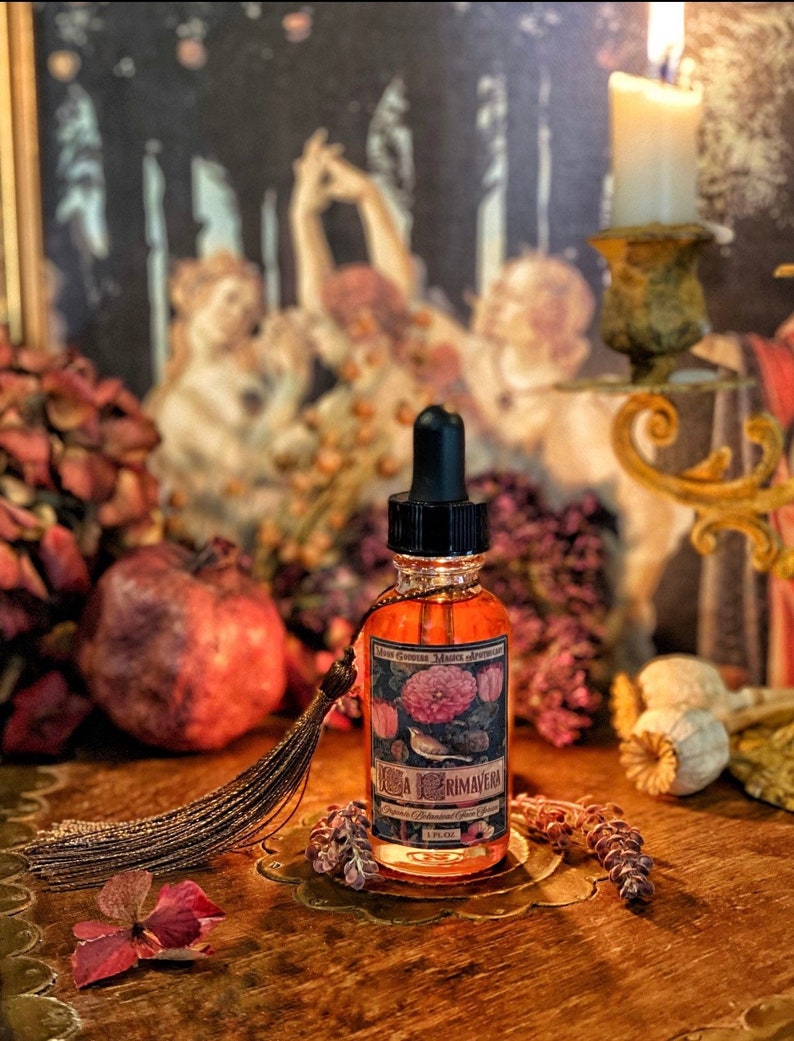 May include: A small amber glass bottle with a black dropper top and a label that reads "Rosa Primavera" in a vintage style font. The bottle is sitting on a wooden surface with dried flowers and a tassel.