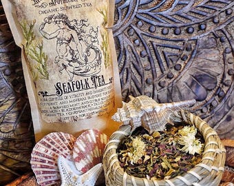 May include: An image featuring a package of "SeaFolk Tea" with a mermaid design, alongside seashells, a starfish, and a woven basket filled with dried herbs. The tea package is sealed with a red wax stamp, set against a carved wooden background.