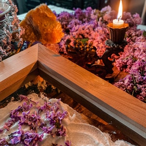 May include: A wooden frame sits on a table with a white surface. Lilac flowers are scattered on the surface. A lit candle sits in a metal candlestick on a bed of lilac flowers.