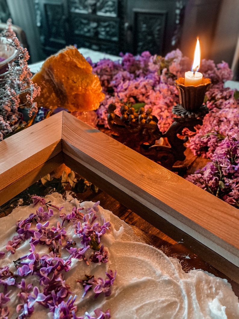 May include: A wooden frame sits on a table with a white surface. Lilac flowers are scattered on the surface. A lit candle sits in a metal candlestick on a bed of lilac flowers.