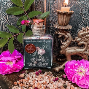 May include: A glass bottle filled with pink Himalayan salt and dried rose petals. The bottle has a black label that reads "Enchanted Rose Garden Rose Otto Ritual Bathing Salts".