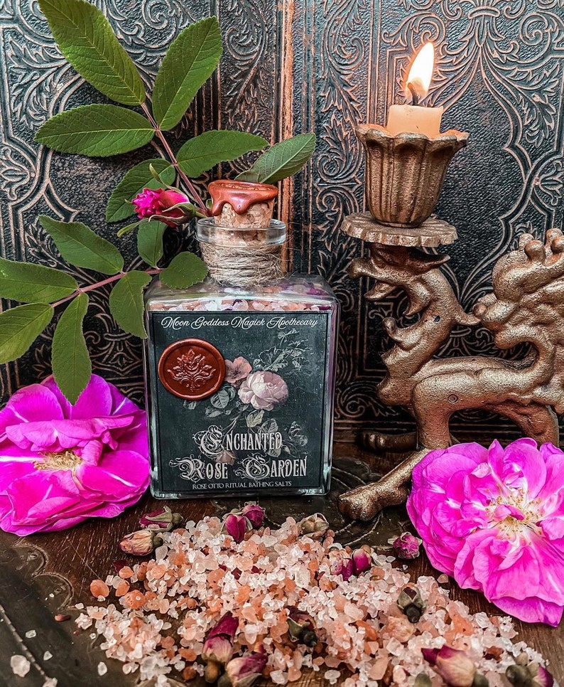 May include: A glass bottle filled with pink Himalayan salt and dried rose petals. The bottle has a black label that reads "Enchanted Rose Garden Rose Otto Ritual Bathing Salts".