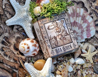 May include: A small glass bottle with a copper top and a label that reads "Moon Goddess Magick Sea Siren". The bottle is surrounded by seashells, starfish, and moss.