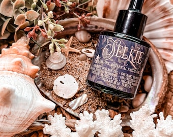 May include: A dark glass bottle with a dropper top, labelled "Selkie Organic Botanical Facial Oil". The bottle is surrounded by seashells and coral on a woven surface.