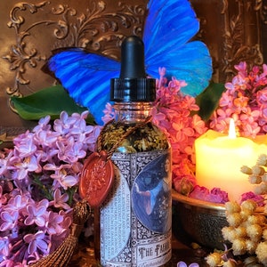 May include: A glass bottle with a dropper top filled with a clear liquid and herbs. The bottle has a label with the text "The Faerie" and "Enchanted Garden". The bottle is surrounded by purple flowers and a blue butterfly.