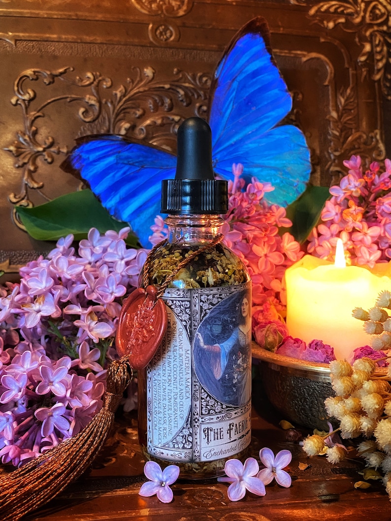 May include: A glass bottle with a dropper top filled with a clear liquid and herbs. The bottle has a label with the text "The Faerie" and "Enchanted Garden". The bottle is surrounded by purple flowers and a blue butterfly.