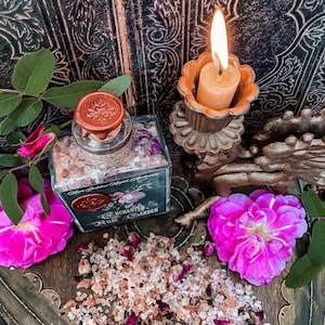 May include: A glass jar filled with pink Himalayan salt and dried rose petals. The jar has a black label that reads "Enchanted Rose Garden" and a wax seal with a rose design. The jar is sitting on a wooden surface with a lit candle and pink roses.