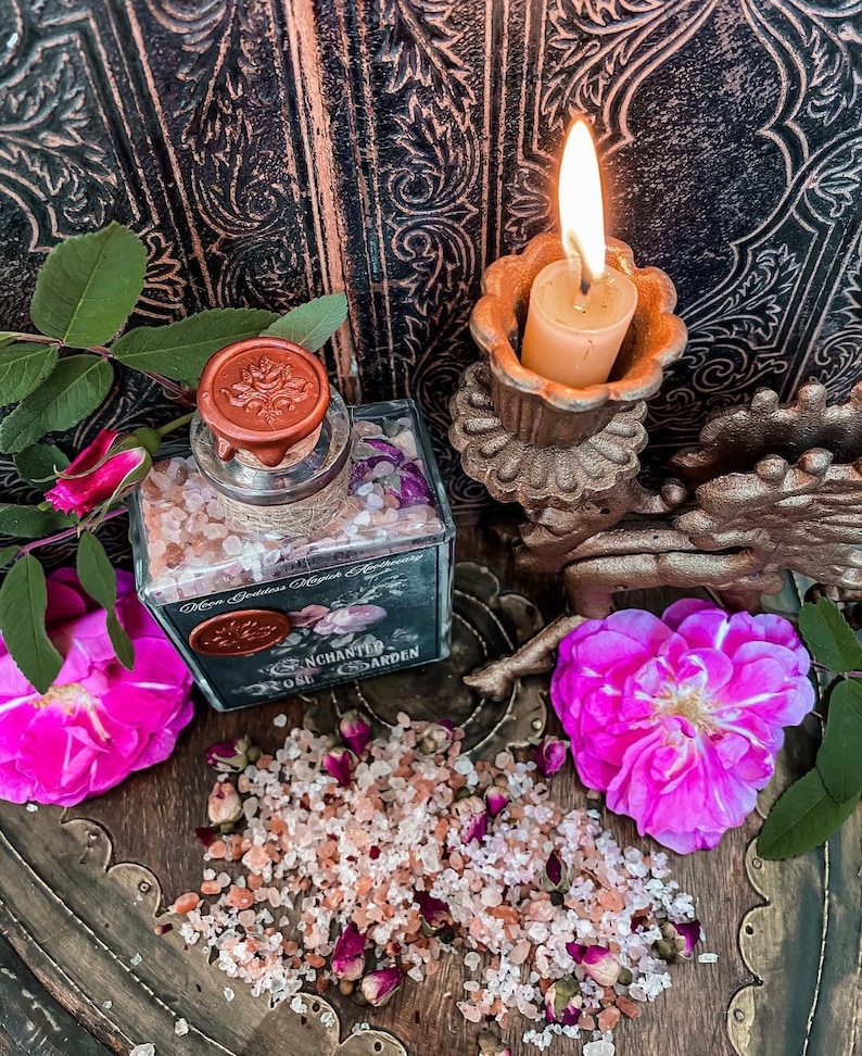 May include: A glass jar filled with pink Himalayan salt and dried rose petals. The jar has a black label that reads "Enchanted Rose Garden" and a wax seal with a rose design. The jar is sitting on a wooden surface with a lit candle and pink roses.