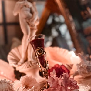 May include: A decorative display featuring a small, ornate glass with a dark red liquid, set on a seashell. A small statue and pink flowers are also present. The glass has gold accents and is surrounded by other seashells.