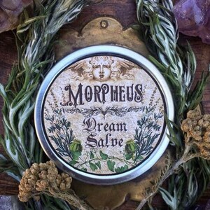 May include: A silver tin with a vintage label that reads "Morpheus Dream Salve". The label features a stylised image of a woman's face with wings and floral designs.