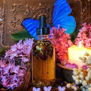 May include: A glass bottle with a dropper filled with a golden liquid and dried flowers. The bottle is surrounded by purple lilac flowers, a blue butterfly, and a white candle.
