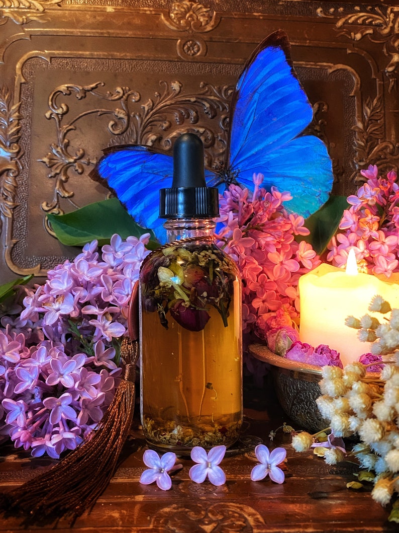 May include: A glass bottle with a dropper filled with a golden liquid and dried flowers. The bottle is surrounded by purple lilac flowers, a blue butterfly, and a white candle.
