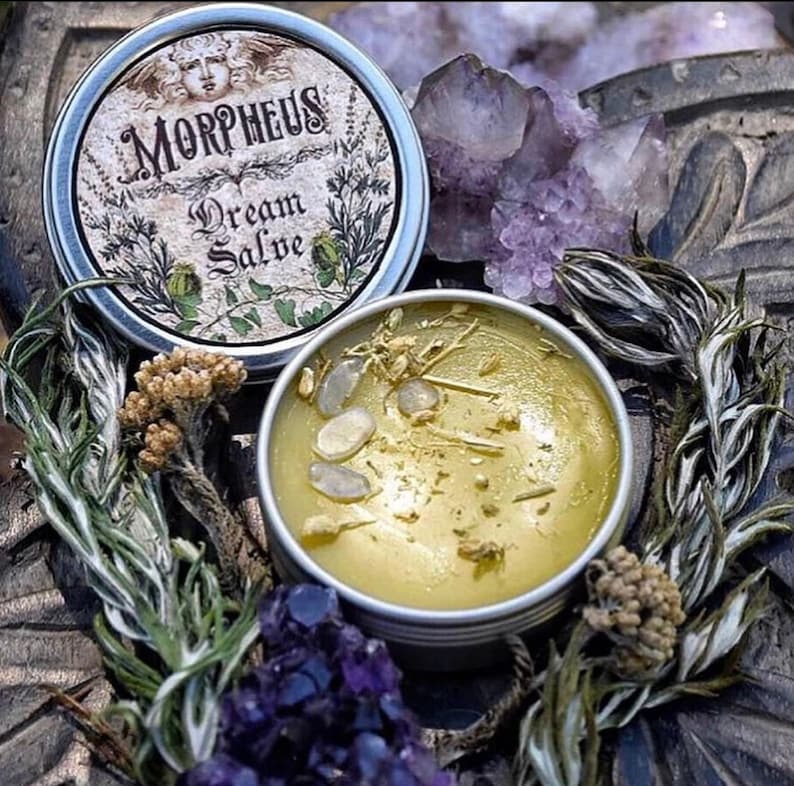 May include: A small tin container labelled "Morpheus Dream Salve". The container is filled with a yellow salve and contains small white crystals and herbs. The container is surrounded by green plants and purple crystals.