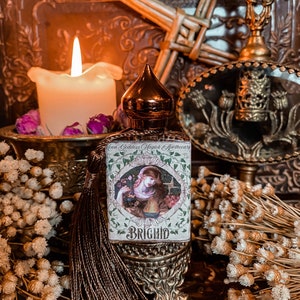 May include: A small bottle of perfume with a gold cap and a label that says "Green Goddess Magick Apothecary" and "Brigid". The bottle is sitting on a decorative stand with a tassel.